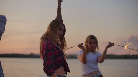 Two-girls-are-dancing-with-big-bengal-lights-on-the-river-coast.-This-is-cool-theamy-summer-evening-on-the-open-air-party-at-pink-sunset.
